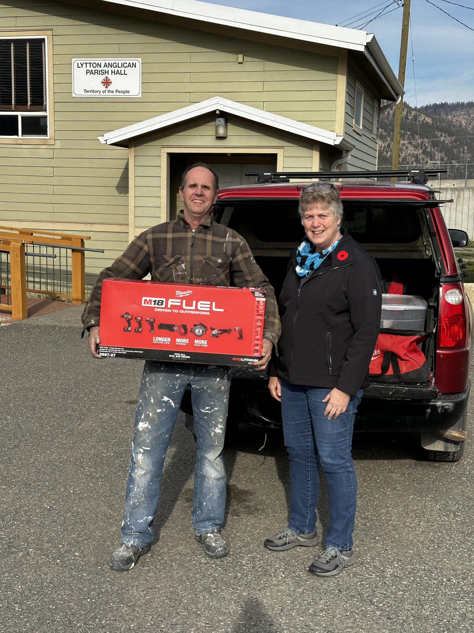 A man holding a Milwaukee toolset box while standing beside a woman in front of Lytton Anglican Parish Hall.