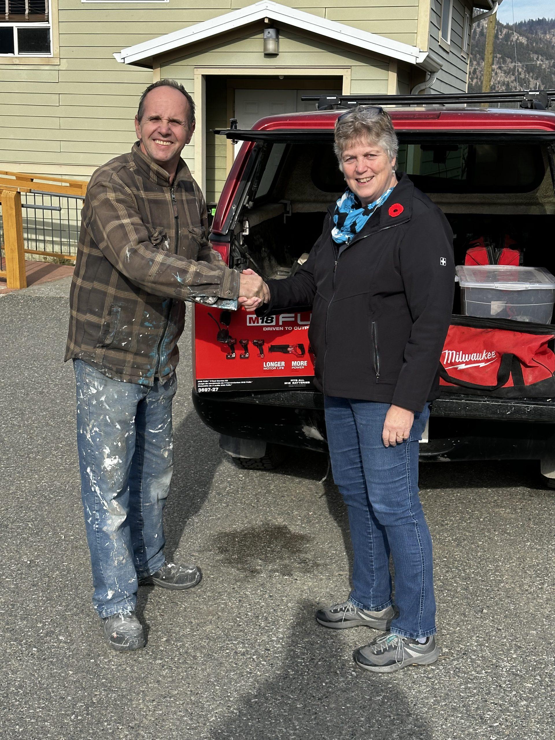 A man and a woman shaking hands beside a vehicle in front of Lytton Anglican Parish Hall.